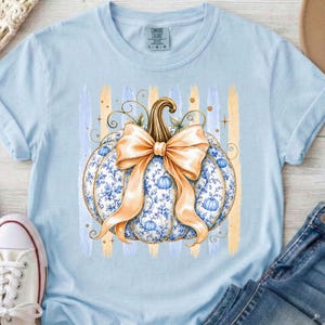 May include: A light blue t-shirt featuring a central graphic of a pumpkin with a blue and white floral pattern, adorned with a large tan ribbon bow. The design is set against a background of vertical light blue and tan brushstroke stripes.