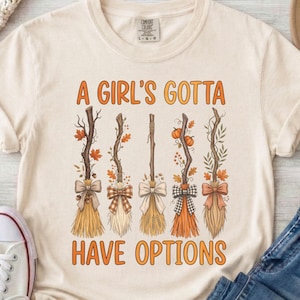May include: An ivory short-sleeve t-shirt featuring a graphic of five stylized brooms decorated with autumn leaves, pumpkins, and bows. The text "A GIRL'S GOTTA HAVE OPTIONS" is printed in orange above and below the broom illustrations.