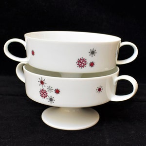 Peut inclure: Deux bols à soupe en céramique blanche avec des poignées, empilés sur un socle. Chaque bol présente un motif de flocon de neige rouge et gris. Les bols sont conçus pour servir de la soupe ou d'autres plats.