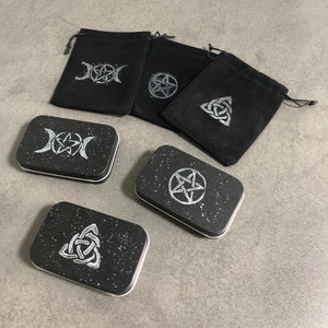 May include: Four small black velvet pouches with silver-colored symbols and four small black tins with silver-colored symbols on a gray surface. The symbols are a triple moon, a pentagram, a triquetra, and a pentagram.