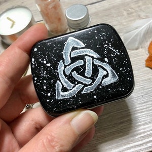 May include: A black metal tin with a white Celtic knot design on the lid. The tin has a speckled, galaxy-like background.