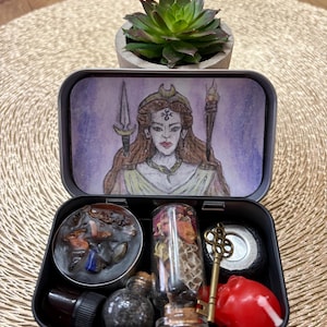 Può includere: Una scatola di metallo nera con un'illustrazione ad acquerello di una donna con una corona e una lancia. All'interno ci sono piccole bottiglie, una chiave, un cuore rosso e una piccola pianta succulenta in un vaso di cemento.