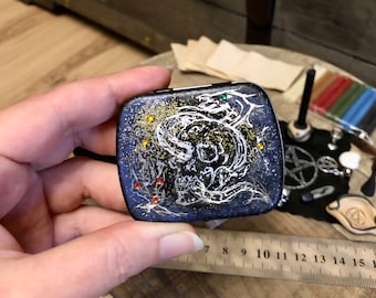 Può includere: Una piccola scatola quadrata nera con uno sfondo di glitter dorati e un disegno a linea bianca di uno scheletro con un design a spirale. La scatola ha alcuni piccoli punti colorati intorno allo scheletro.