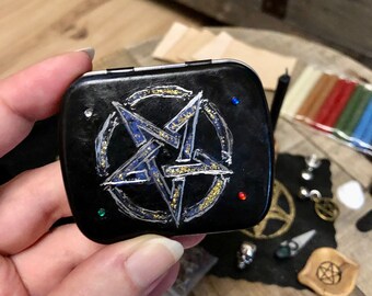 Può includere: Una piccola scatola di metallo nero con un design a pentagramma dorato. La scatola ha un bordo argentato ed è decorata con piccole pietre preziose rosse e verdi.