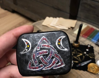 Può includere: Una scatola quadrata nera con un design a nodo celtico argentato, rosso e blu. Il nodo è circondato da due lune crescenti argentate con accenti arancioni e gialli.