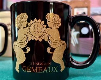 Pode incluir: Caneca de cerâmica preta com uma ilustração dourada de duas figuras segurando um sol. A caneca tem uma alça preta e o texto "21 MAI-20 JUIN GEMEAUX" em ouro e verde. A caneca é um design do zodíaco de Gêmeos.
