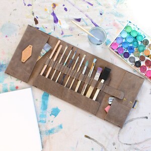 May include: A brown leather roll-up case with a flap closure and a snap button. The case is open and contains a variety of paintbrushes, including round, flat, and angled brushes. The case is on a white surface with paint splatters.