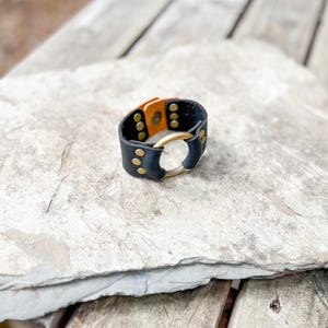 May include: A black leather bracelet with a gold-colored metal ring and rivets. The interior of the bracelet is tan. The bracelet is resting on a light gray stone surface.