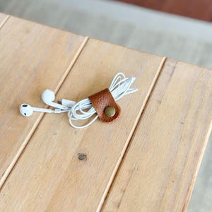 May include: A brown leather cable organizer with a brass snap button holds white earbud wires. White earbuds are placed to the left of the organizer. The organizer is on a light brown wooden surface.