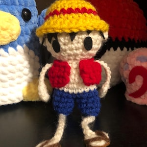 Op de afbeelding: Gehaakte Luffy-pop met een gele hoed met rode rand, een wit shirt, rode handschoenen en een blauwe korte broek. De pop heeft zwarte ogen en bruine sandalen. Andere knuffels op de achtergrond.