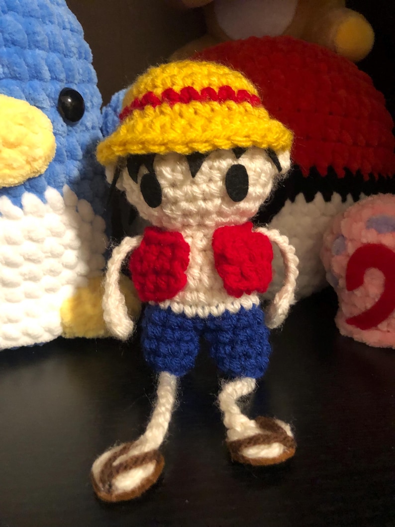 Puede incluir: Mu&ntilde;eco de Luffy tejido a ganchillo, con un sombrero amarillo con ribete rojo, una camisa blanca, guantes rojos y pantalones cortos azules. El mu&ntilde;eco tiene ojos negros y sandalias marrones. Otros peluches en el fondo.