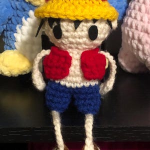 Puede incluir: Mu&ntilde;eco de Luffy tejido a crochet, con sombrero de paja amarillo con banda roja, chaleco rojo, pantalones cortos azules y cuerpo blanco. El mu&ntilde;eco tiene ojos negros y sandalias marrones. El amigurumi hecho a mano es un personaje de anime.