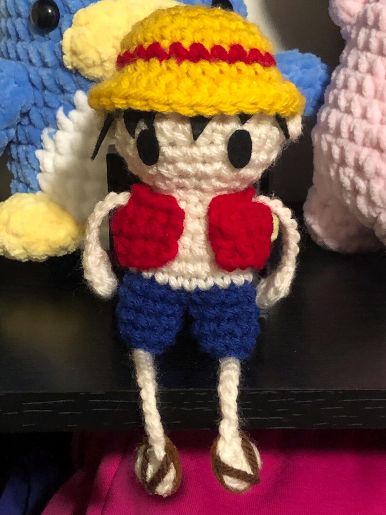 Puede incluir: Mu&ntilde;eco de Luffy tejido a crochet, con sombrero de paja amarillo con banda roja, chaleco rojo, pantalones cortos azules y cuerpo blanco. El mu&ntilde;eco tiene ojos negros y sandalias marrones. El amigurumi hecho a mano es un personaje de anime.