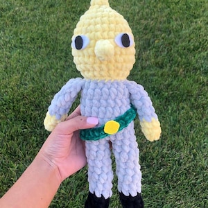 Puede incluir: Muñeca de ganchillo hecha a mano con cabeza amarilla y cuerpo gris. La muñeca tiene ojos grandes y redondos, una nariz pequeña y un cinturón verde con hebilla amarilla. Los pies de la muñeca son negros.
