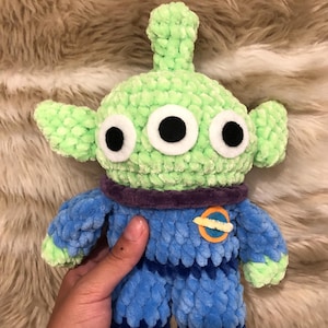Può includere: Un peluche all'uncinetto a forma di alieno a tre occhi con testa verde chiaro, orecchie a punta e una piccola antenna. Il corpo è blu con un colletto viola scuro e una toppa circolare arancione e blu a forma di pianeta sul petto.
