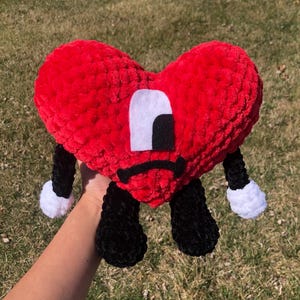 Könnte beinhalten: Ein rotes, herzförmiges Plüschtier mit schwarzen Beinen und Armen sowie weißen Akzenten. Das Spielzeug hat ein weißes und schwarzes Augendesign und einen traurigen Mund. Das Spielzeug besteht aus einem weichen, strukturierten Material.