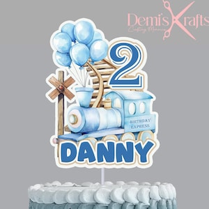 Puede incluir: Un pastel de dos niveles con glaseado azul degradado, coronado con un adorno de pastel de tren azul. El adorno presenta un tren, globos, el número 2 y el nombre "DANNY". El pastel está sobre un soporte blanco.