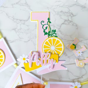 May include: A pink and yellow cake topper for a first birthday, featuring the number one, a lemon slice, and the name "Polly". The topper has a clear plastic stick. A banner with the letter "E" and daisy decorations is in the background.