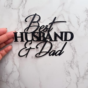 以下が含まれることがあります： 「Best Husband & Dad」の文字が書かれた黒いグリッターケーキトッパー。筆記体とブロック体のフォントを使用。白とグレーの大理石の背景に、手で持たれています。
