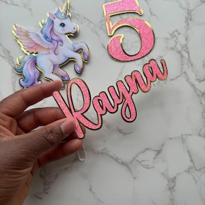 Pode incluir: Um conjunto de topos de bolo de aniversário com um unicórnio com detalhes dourados, o número 5 e o nome "Rayna" em glitter rosa com bordas douradas. O unicórnio tem um corpo branco, olhos azuis e uma crina e cauda arco-íris.
