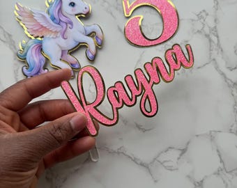 May include: A collection of birthday cake toppers featuring a unicorn with gold accents, the number 5, and the name "Rayna" in pink glitter with gold trim. The unicorn has a white body, blue eyes, and a rainbow mane and tail.