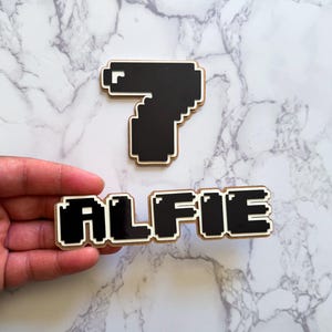 Può includere: Due ritagli di legno pixelati: il numero 7 e il nome "ALFIE". Il numero 7 è nero con un contorno bianco. Il nome "ALFIE" è anch'esso nero con un contorno bianco.