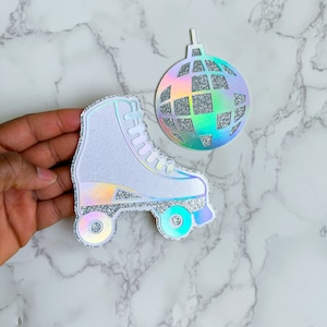 May include: A white roller skate and a disco ball ornament with iridescent and glitter accents. The roller skate has silver glitter trim and wheels. The disco ball ornament has a silver glitter centre and a holographic rainbow effect.