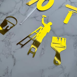 May include: Gold-colored cake toppers and decorations, including a number 70, a painter on a ladder, a paint bucket, and a paintbrush. The items are arranged on a marble-patterned surface.