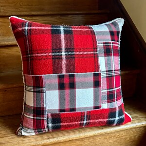 May include: Red and white plaid throw pillow with a patchwork design. The pillow has a white sherpa trim around the edges.