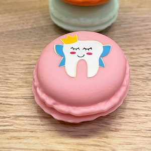 May include: A stack of four colorful macarons with a pink macaron on the bottom featuring a cartoon tooth with a crown and wings.