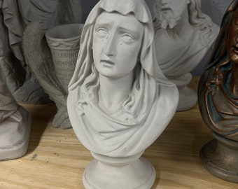 May include: White plaster bust sculpture of a woman with a sorrowful expression. She is wearing a draped robe and has her head slightly tilted down. The sculpture is on a white pedestal.