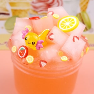May include: A clear plastic container filled with pink slime, topped with white cubes, strawberry and lemon slices, and a yellow axolotl charm.