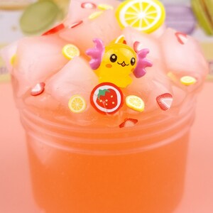May include: Pink slime with a yellow cartoon character, strawberries, and lemon slices. The slime is in a clear plastic container.