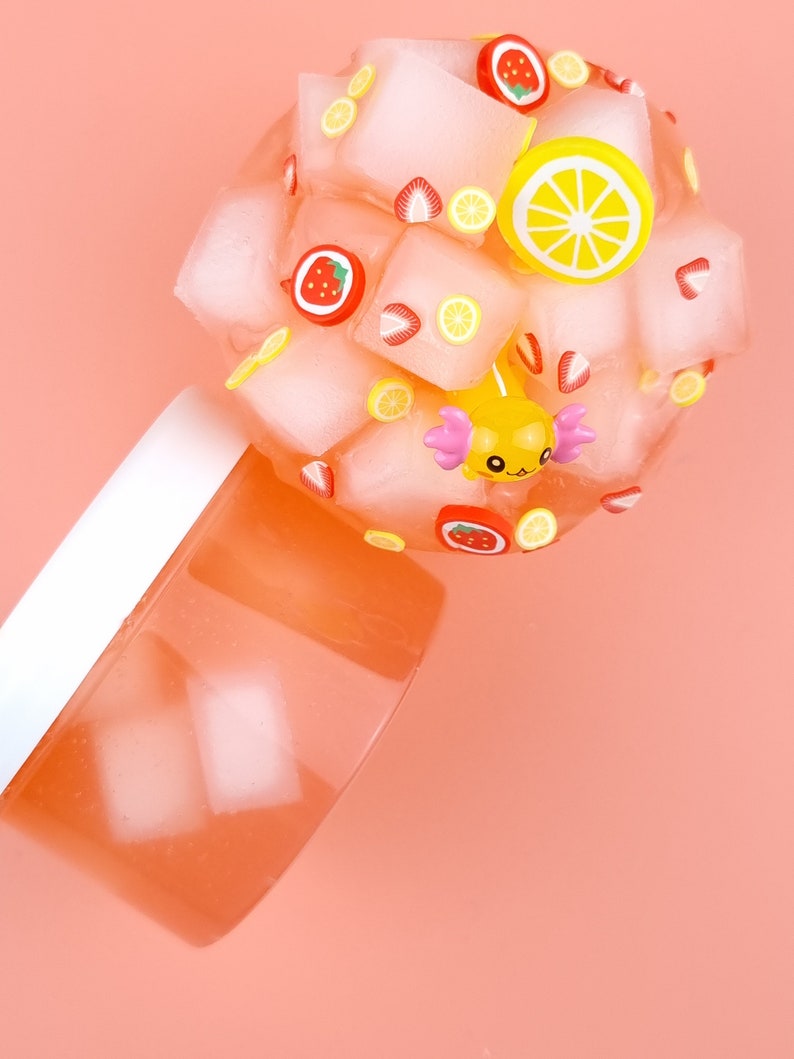 May include: A pink slime with ice cubes, lemons, and strawberries. The slime is in a clear container with a white lid.