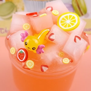 May include: A clear plastic container filled with a pink slime-like substance. The slime is topped with various colorful decorations, including a yellow axolotl figurine, red and yellow strawberry slices, and yellow lemon slices.