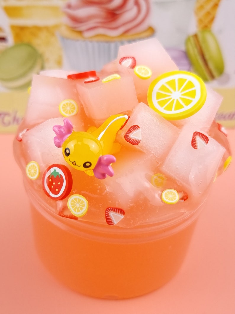 May include: A clear plastic container filled with a pink slime-like substance. The slime is topped with various colorful decorations, including a yellow axolotl figurine, red and yellow strawberry slices, and yellow lemon slices.