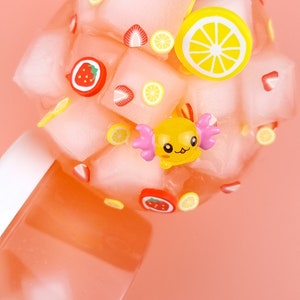 May include: A pink and white slime with a yellow cartoon character, strawberries, and lemons. The slime is in a clear container.