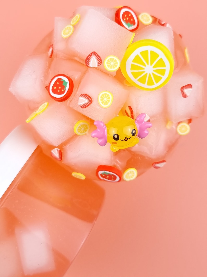 May include: A pink and white slime with a yellow cartoon character, strawberries, and lemons. The slime is in a clear container.