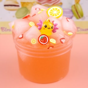 May include: A pink and orange slime with a yellow cartoon character, strawberries, and lemon slices. The slime is in a clear plastic container.