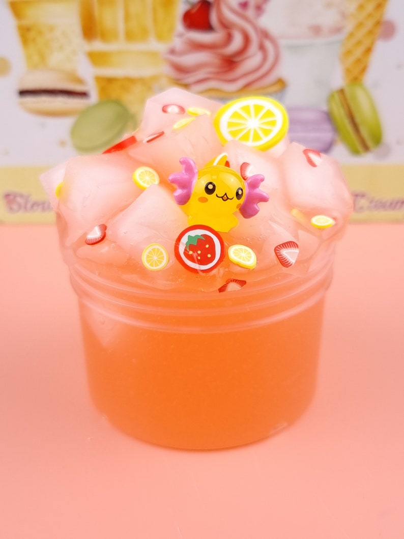 May include: A pink and orange slime with a yellow cartoon character, strawberries, and lemon slices. The slime is in a clear plastic container.