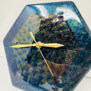 May include: A geometric-shaped clock with a dichroic glass face. The clock features a gold-colored clock mechanism and hands. The glass has a blue and iridescent color scheme, with hints of gold, green, and purple. The clock is a decorative item.