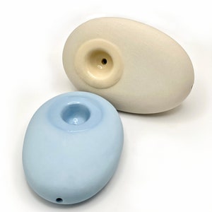 May include: Two ceramic pipes shaped like pebbles. One is light blue and the other is off-white.