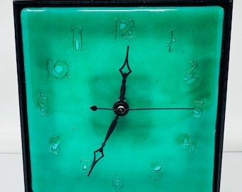May include: Square clock with a teal face and black frame. The clock face features stylized numbers and black hands. The clock's face has a textured appearance. The clock is a decorative item.