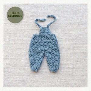 May include: Blue crocheted baby overalls with adjustable straps. The overalls are laid flat on a white textured surface. The text "HÄKEL KLEIDUNG" is visible in the upper left corner of the image.