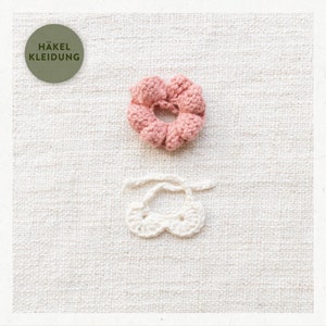 May include: Two crocheted hair scrunchies, one pink and one white. The pink scrunchie is in the shape of a flower. The white scrunchie is in the shape of a bow. The scrunchies are on a white linen background.