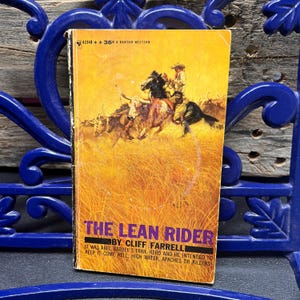 May include: A vintage paperback book titled "The Lean Rider" by Cliff Farrell. The cover features a Western scene with cowboys on horseback and cattle in a field of tall grass. The book is a Bantam Western.