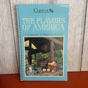 May include: A spiral-bound cookbook titled "The Flavors of America" with a light blue cover. The cover features the "Current" logo and a photograph of a well, baskets of fruit, and the text "A Collection of Regional Recipes".