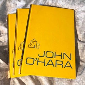 May include: Three hardback books with bright yellow covers. The title "JOHN O'HARA" is printed in navy blue, along with a small house icon. The books are stacked on a textured, light grey surface.
