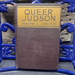 Könnte beinhalten: Ein Vintage-Hardcover-Buch mit dem Titel "Queer Judson" von Joseph C. Lincoln. Das Buch hat einen braunen Einband mit einem goldenen Titelfeld. Titel und Autorenname sind in Schwarz gedruckt. Das Buch liegt auf einem blauen Zierstuh