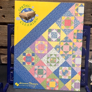 May include: A book titled "Spring Cleaning" with a yellow cover and a blue diagonal border. The cover features a colorful patchwork quilt design with squares in various pastel colours. The Atkinson Designs logo is visible.